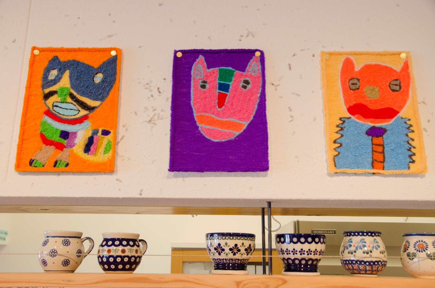鮮やかな色彩で動物の顔が描かれ作品が壁に3枚並べて飾られており、その下にはかわいらしい模様の陶器のマグカップが並んでいるギャラリースペースの写真