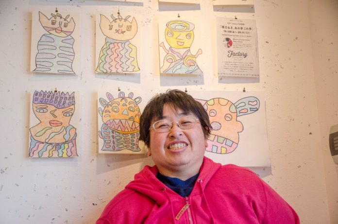 壁一面に展示されたカラフルでユニークなアート作品の前で、笑顔で写っている前川さんの写真