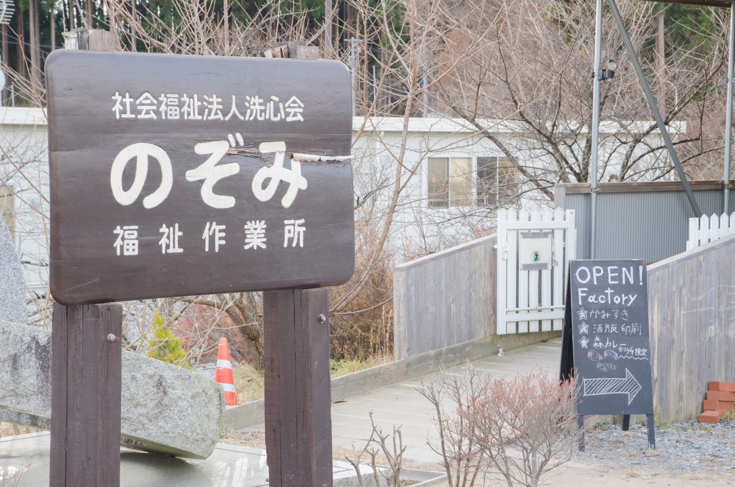 「社会福祉法人洗心会 のぞみ福祉作業所」と書かれた木の看板と、「OPEN! Factory」と書かれた黒板の案内板が立ち、工房の入り口へと続く木製スロープがある風景の写真
