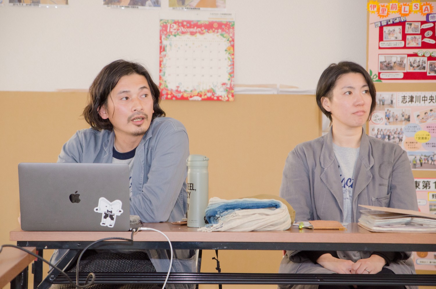 会議室のような場所で、ノートパソコンの前で真剣な表情で話す前川雄一さんと、その横に座る前川あきこさんが写っている写真