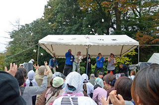 屋外ステージの上から男性たちが景品の入った小袋を観客に向けて投げており、観客たちが手を伸ばしてそれを受け取ろうとしている写真