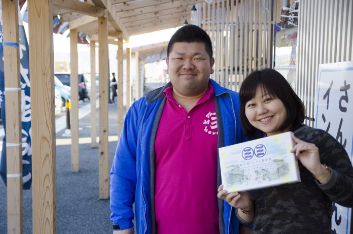 ピンクのポロシャツに青い上着を着た佐藤さんと、女性リポーターが、商店街の軒下で箱入りの商品を持って並んで立っている写真