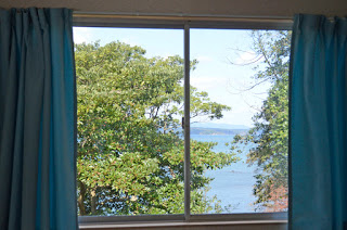 青いカーテンに囲まれた大きな窓から、緑の木々とその向こうに広がる青い海と空が見える風景の写真