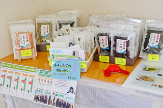 海藻類や乾物の袋詰め商品が棚の上に並び、それぞれに手書き風の値札が付けられ、商品説明のチラシも置かれている販売コーナーを写した写真
