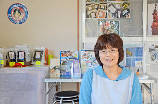 壁際に海藻などの袋詰め商品と観光パンフレットが並べられたテーブルの前で、水色のエプロンを着た中年の女性が笑顔で立っている写真