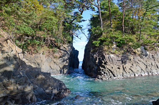 神の力で割れたという伝説が残る、岩が二つに裂けてできた細い海の割れ目を正面から捉え、岩肌とその上に生える木々が見える神割崎の写真