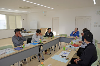 白く明るい会議室で、男性たちが長机を囲んで資料や飲み物を前にしながら話し合っている写真