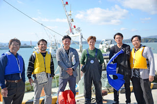 港に停泊中の漁船の前で、ライフジャケットや作業着姿の男性たちが笑顔で並んでいる写真