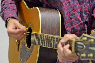 赤いチェックのシャツを着た人物がアコースティックギターを弾いている演奏中の手元の写真