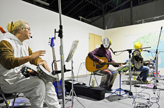 着ぐるみを着たり、アニメのキャラクターのお面をつけた3人が打楽器やギターを手に椅子に座り、屋内の会場で演奏をしている写真