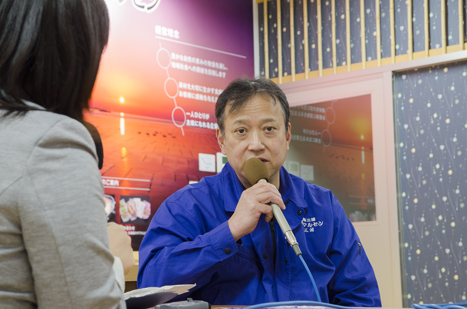 青い作業服を着た三浦さんがマイクを持ち、女性リポーターと向かい合って座りながらインタビューを受けている写真
