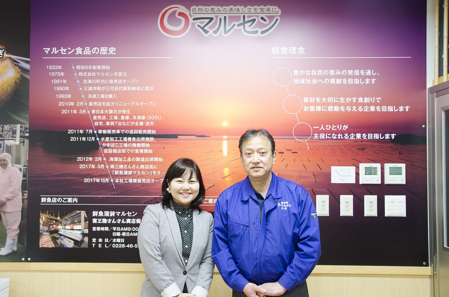 マルセン食品のロゴと歴史、経営理念などが記された展示パネルの前で、グレーのジャケットを着た女性リポーターと青い作業服を着た三浦さんが並んで立ち、カメラに向かって笑顔を見せている写真