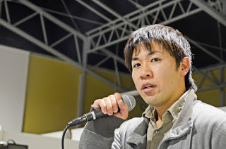 グレーのジャケットを着た男性がマイクを持ち、話をしている場面の写真