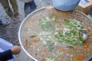 大鍋いっぱいに牛肉やネギ、人参などの具材が煮込まれている芋煮が、屋外で湯気を立てている様子の写真