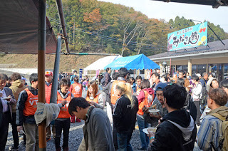 会場前に多くの人が集まり、オレンジ色のボランティアビブスを着た人や食事を手にした来場者が談笑している様子の写真