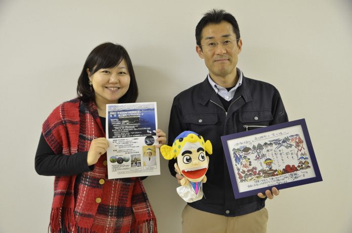 女性リポーターがチラシを持ち、隣の阿部さんがキャラクターの人形と色鮮やかなイラストの額を持って並んで立っている写真
