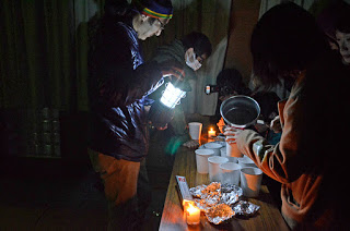 暗い室内で停電時を想定してランタンやロウソクの明かりのもと、数人が紙コップにスープを注いだり、アルミホイルに包まれた食べ物を準備している様子の写真