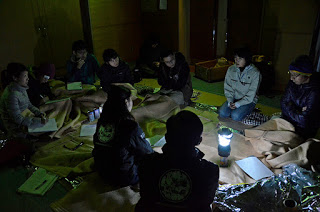 薄暗い室内で防災訓練に参加している十人ほどの人々が毛布や銀色の保温シートを使って床に座り、ランタンの光の中で真剣に話し合っている写真