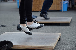 正方形の木製のステージの上で二人の人物がタップダンスをしている様子が写されており、片方の人物は黒と白のタップシューズを履いてジャンプしている瞬間が捉えられ、もう一人は黒いタップシューズを履いて踊っている写真