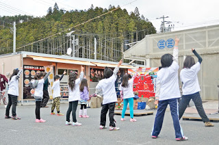 背景には看板やポスターが貼られた建物があり、木々、電線が見える屋外の広場で白いシャツを着た子どもたちが右腕を上げてダンスを踊っている後ろ姿の写真