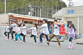 背景には建物や木々があり、屋外イベントで子どもたちが一列に並び、片足を上げて腕を伸ばしダンスを踊っている様子が写された写真
