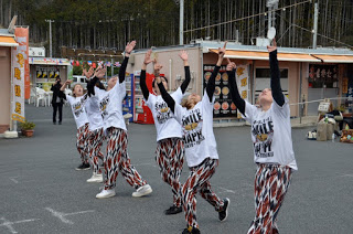 背景には看板や装飾のある小さな建物や屋台が並ぶ屋外のイベント会場で、「SMILE HAPPY」と書かれた白いTシャツと柄のパンツを着た6人の人物が、横一列に並び揃ってダンスを踊っている様子が写された写真