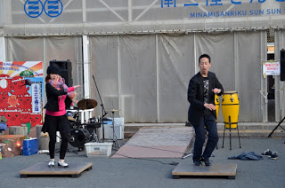 背後にはドラムセットや音楽機材がある屋外のステージの上で、二人の人物が立ってダンスをしている様子が写された写真