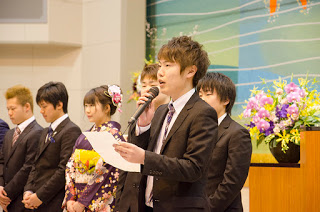 演台花が設置された演台の前で、振り袖姿の女性やスーツ姿の男性の新成人が一列に並び、そのうちの一人がマイクを持って紙を読み上げている様子を写した写真