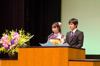 左側に花が生けられた木製の演台に立つ男性と女性の新成人の代表が成人の抱負を語っている様子の写真