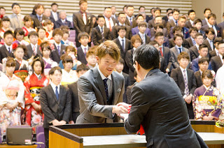 背景には振袖姿やフォーマルなスーツを着た男性が列になって立っている多くの新成人が出席する式典の場で、前方にいる男性が演台越しに町長と握手を交わしている様子の写真