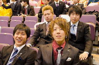 紫色の座席が並ぶ講堂で、ネクタイをつけスーツや着た新成人の4人の男性が座っている様子が写されている写真