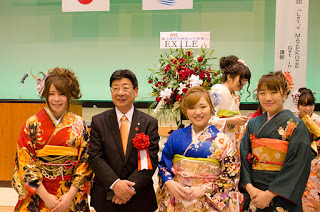 国旗と町旗が掲げられ、EXILEから送られてきた赤と白の祝花が飾られた前で、中央に赤白のリボンを胸につけたスーツ姿の町長を挟んで着物を着た3人の女性が並んでいる記念写真
