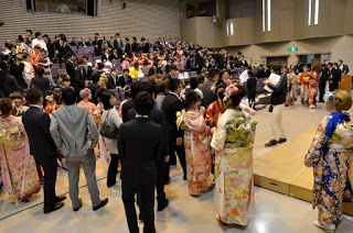 フォーマルな装いをした男性や、着物を着た女性たちがホールのような場所に集まっている成人式の様子の写真