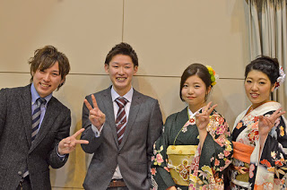 スーツを着た2人の男性と、振り袖姿の2人の女性が並んで立ち、全員がピースサインをしてポーズをとっている様子を写した写真