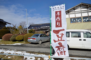 雪の残る道路沿いに「手作り豆腐」と書かれたのぼりが立っており、その後ろに車が停まった建物が見えている写真