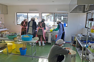 大きな容器や様々な調理器具が置かれた調理場で、4人の女性が窓辺のシンクで作業をし、1人の女性が手前で床に置かれた大きな鍋をしゃがんで混ぜている写真