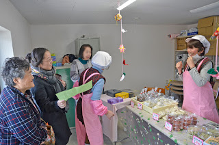 豆腐などの商品がずらっと並んだカウンターの中と外に、ピンク色のエプロンと白い帽子を身に着けた女性が立ち、店を訪れた3人の女性と楽しそうに話をしている写真