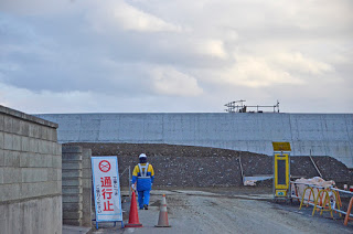 「通行止め」の看板の先を作業員の男性が歩いており、その背後には建設中の防潮堤が見えている写真