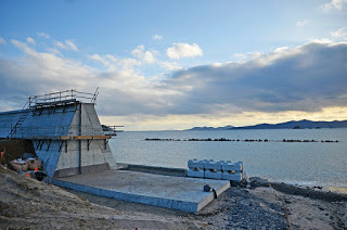 海岸沿いにコンクリートが敷かれ、途中まで建設された防潮堤の後ろに穏やかな海が広がっている写真