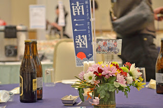 藤色のテーブルクロスが掛けられたテーブルに、写真と小さな南三陸ののぼりが立てられた花が飾られ、ビールの瓶などが準備されている様子の写真