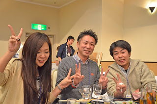 グラスや料理が乗ったテーブルの席に座った女性と2人の男性が笑顔でピースサインをしている写真