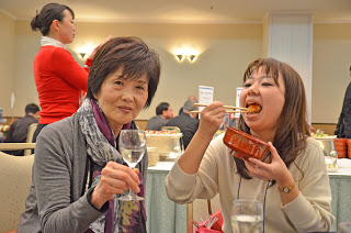 透明の飲み物が入ったワイングラスを掲げている女性と、その横で手に持った丼から料理を口に運んでいる女性の写真