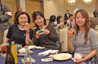 ビールやジュースの瓶が並んだテーブルの席に座っている女性3人が、料理などをこちらに見せながら笑顔で写っている写真