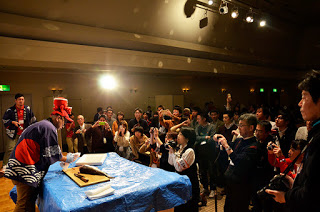 照明を落とした会場で、スポットライトを浴びながら、ブルーシートが敷かれた台の上で大きな魚をさばいている法被を着てタコの人形がついた帽子をかぶった男性と、その周りに集まってカメラや携帯電話を手に撮影をしている人々の写真