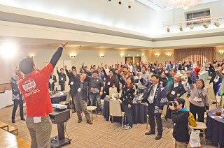 ステージに立った赤いポロシャツの男性に合わせ、会場いっぱいの参加者たちが、立ち上がって右のこぶしを突き上げている写真