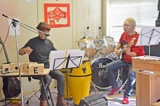 木琴や木の箱でできた楽器、ドラムセットが並ぶ中、ギターを持った男性ともう一人の男性が椅子に座って演奏をしている様子の写真