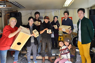 大小の木の箱や木琴を持った男女6人と、子ども連れの男女が集まって写った記念写真