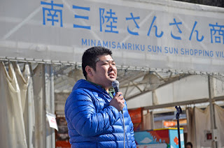 青いダウンジャケットを着た男性がマイクを持って話をしている写真