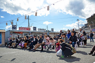 屋外のオープンスペースに並んだ椅子にたくさんの来場者が座って賑わいを見せている商店街の写真