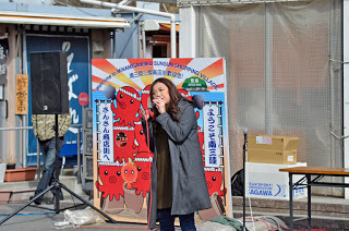 たくさんのタコのイラストが描かれた大きな看板の前でマイクを持った女性が歌っている様子の写真